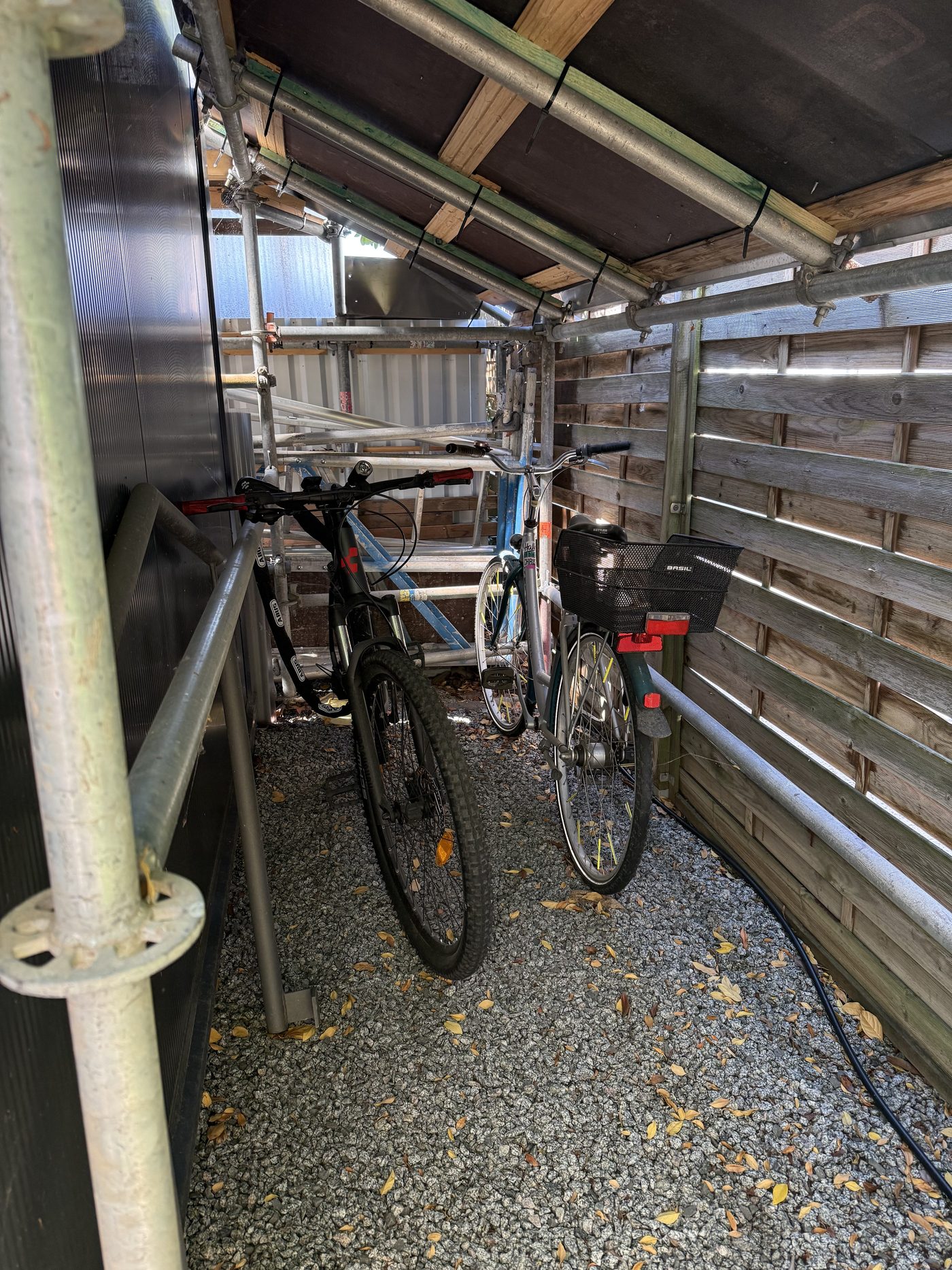 Fahrrad-Unterstand aus Gerüstrohren: Dachlatten quer über den Rohren, darüber lichtdurchlässige Wellplatten, darunter stehen die Räder trocken